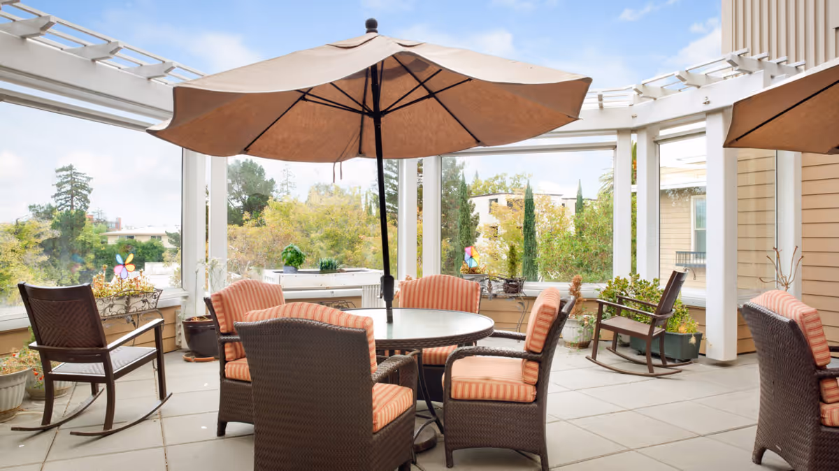 Outdoor patio area with a round glass table surrounded by four cushioned wicker chairs and two wooden rocking chairs. A large beige umbrella is positioned over the table. The patio is enclosed with white framing and glass panels, overlooking trees and nearby buildings under a partly cloudy sky.