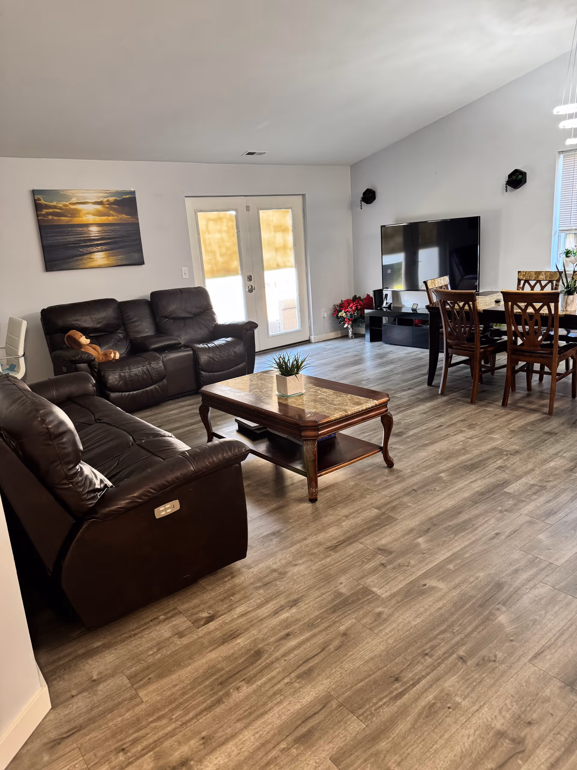 Open-plan living and dining room with brown leather sofas, a wooden coffee table, a wall-mounted TV, dining table and French doors on gray wood-look flooring.