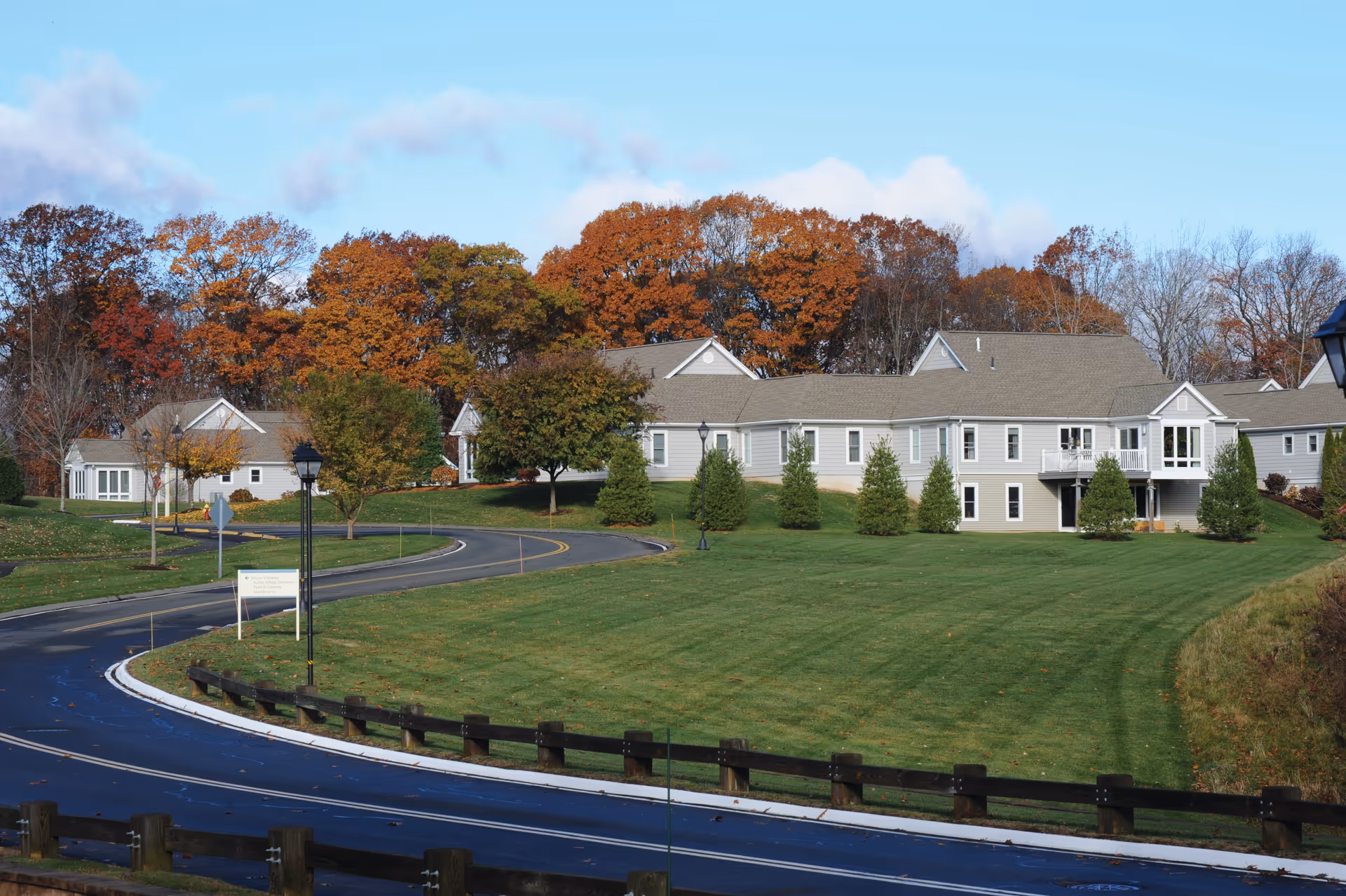 Exterior view of a senior living facility building with beige siding and multiple peaked roofs, surrounded by green lawns, trees with autumn foliage, and a curved paved driveway.
