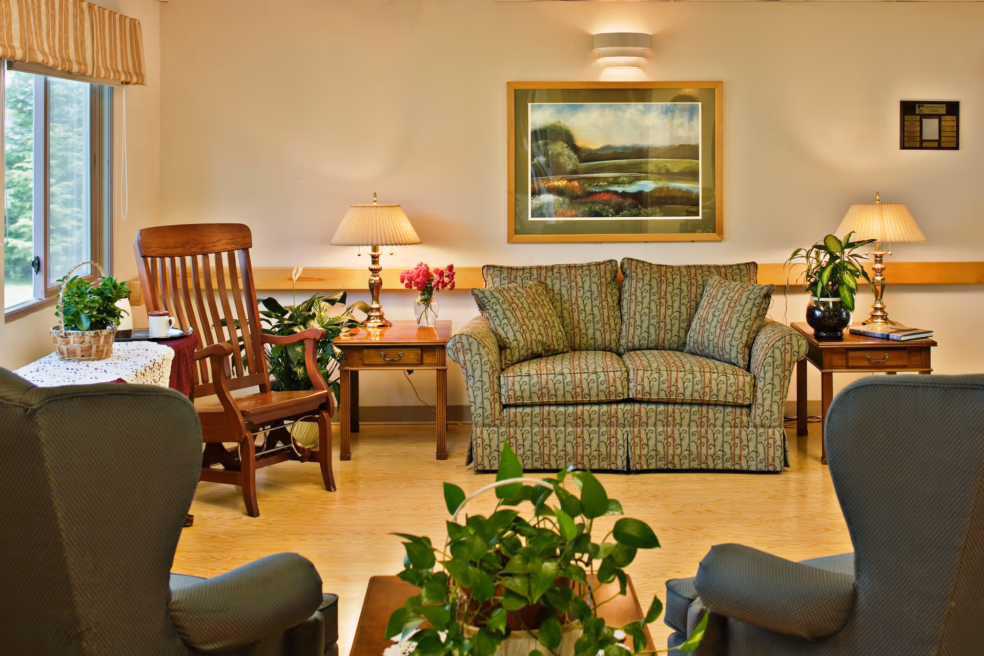 A cozy living room with a patterned sofa, two armchairs, and a wooden rocking chair. There are two side tables with lamps, plants, and flowers. A large window with a striped valance lets in natural light, and a framed landscape painting hangs on the wall above the sofa.