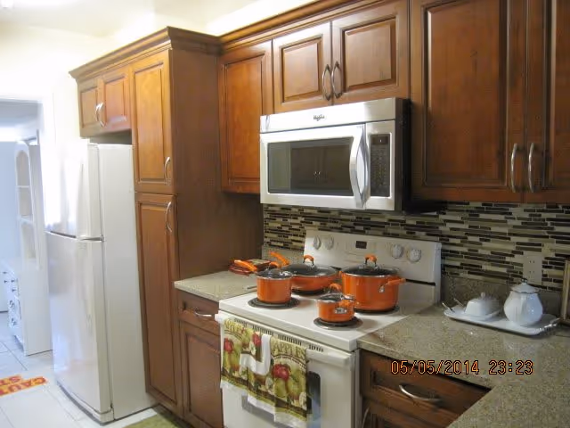 A kitchen area in an assisted living facility featuring wooden cabinets, a white refrigerator, a white stove with orange pots and pans on it, a microwave above the stove, and a countertop with a teapot and cups on a tray. The backsplash has a mosaic tile design.