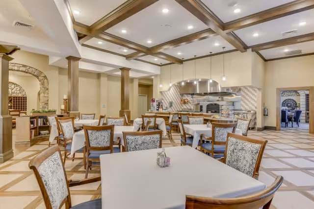 A spacious dining room in a senior living facility with multiple tables covered in white tablecloths and surrounded by wooden chairs with patterned cushions. The room features a coffered ceiling with recessed lighting, a large open kitchen area with a stainless steel oven and range hood, and decorative columns. There is an arched doorway with stone accents leading to another room.