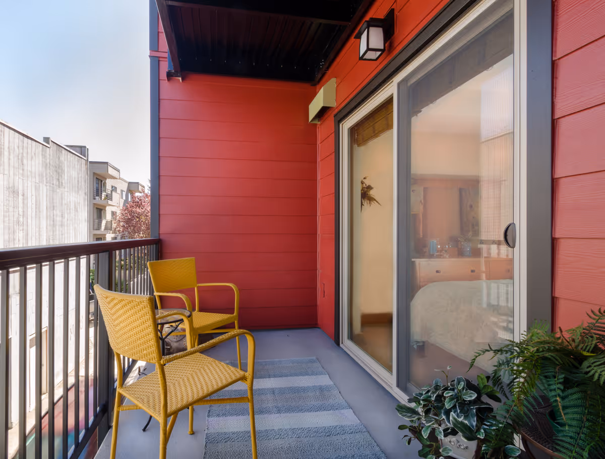 A small balcony with two yellow chairs and a striped rug. The balcony has red walls and a black railing. There are potted plants on the floor near a sliding glass door that leads into a bedroom.