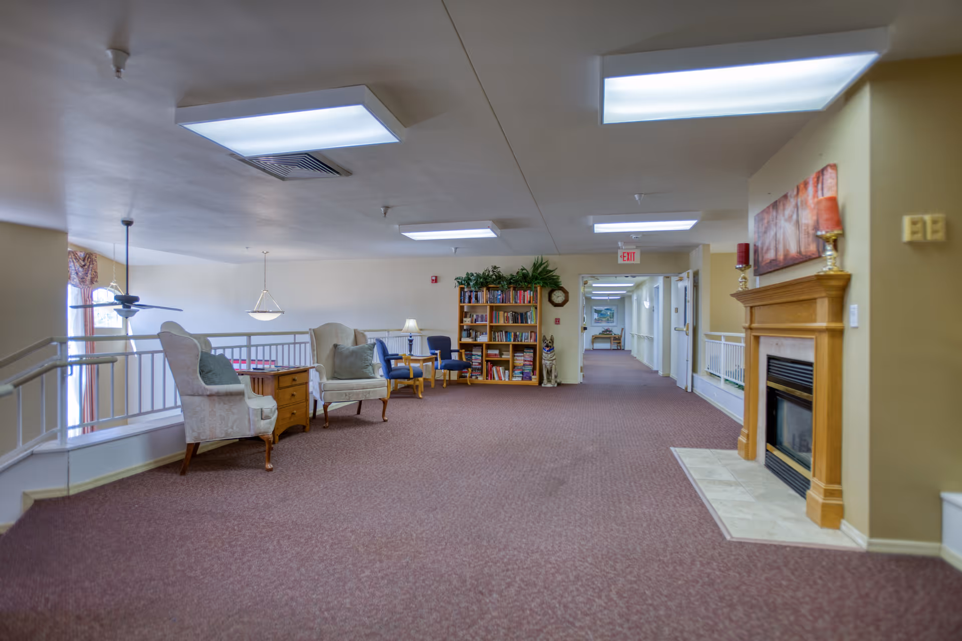 A spacious interior common area with carpeted floor, featuring a wooden fireplace on the right, a bookshelf filled with books and decorative plants at the back, and several upholstered chairs arranged near a small wooden table. The area is well-lit with ceiling lights and has a hallway leading further into the building.
