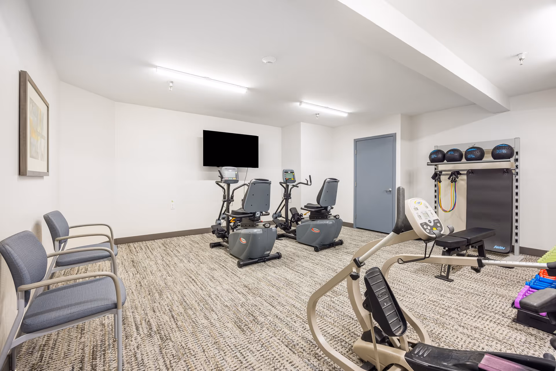 A small fitness room with exercise equipment including two recumbent stationary bikes, a rowing machine, a bench, medicine balls, resistance bands, and dumbbells. There are two gray chairs along the left wall and a flat-screen TV mounted on the wall in front of the exercise bikes. The room has white walls, a gray door, and patterned carpet flooring.