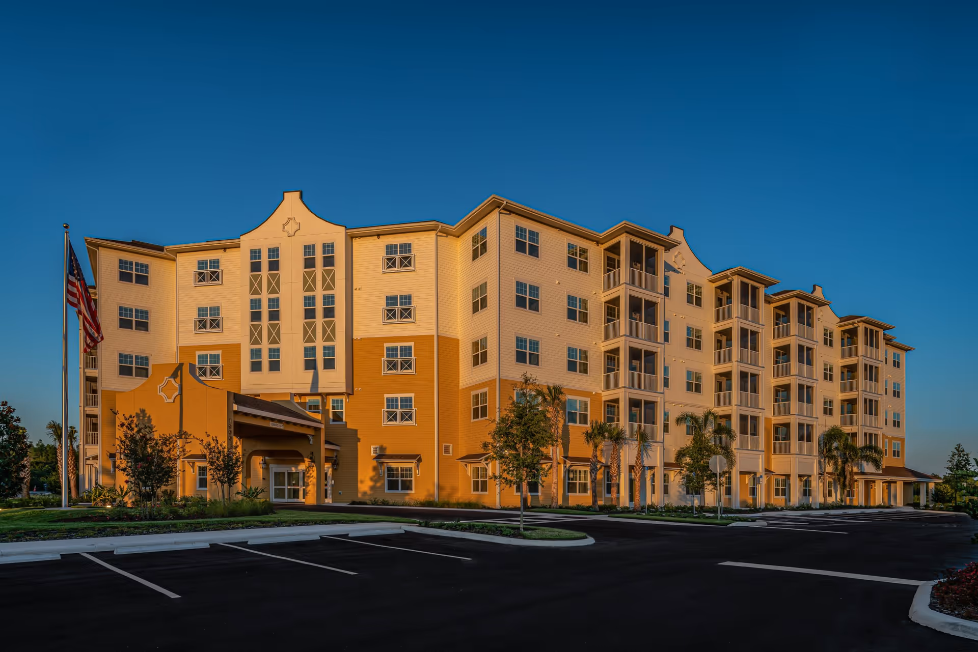 Exterior view of a multi-story senior living facility building named Discovery Village Trinity during sunset, with a clear blue sky, an American flag on a flagpole, palm trees, and an empty parking lot in front.