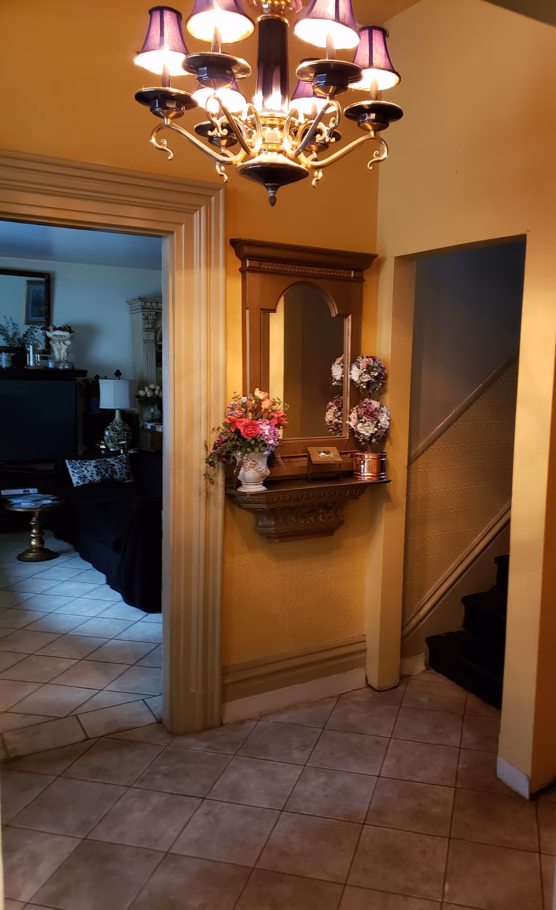 Interior view of a hallway with yellow walls and tiled floor. A decorative wooden wall-mounted mirror with a small shelf below holds a vase of flowers and other small items. To the left, an open doorway leads to a living room with a black couch, a TV, and various decorative items. To the right, a staircase with dark carpeted steps ascends. A chandelier with multiple lights and purple lampshades hangs from the ceiling.