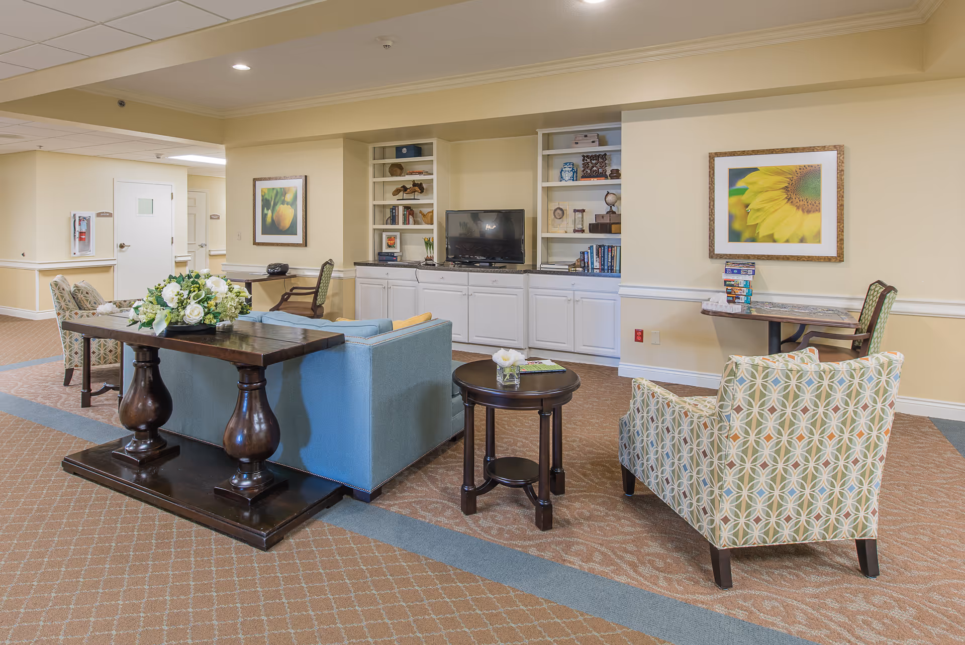 Bright common lounge with sofas and patterned armchairs arranged around tables facing a TV and built-in shelving.