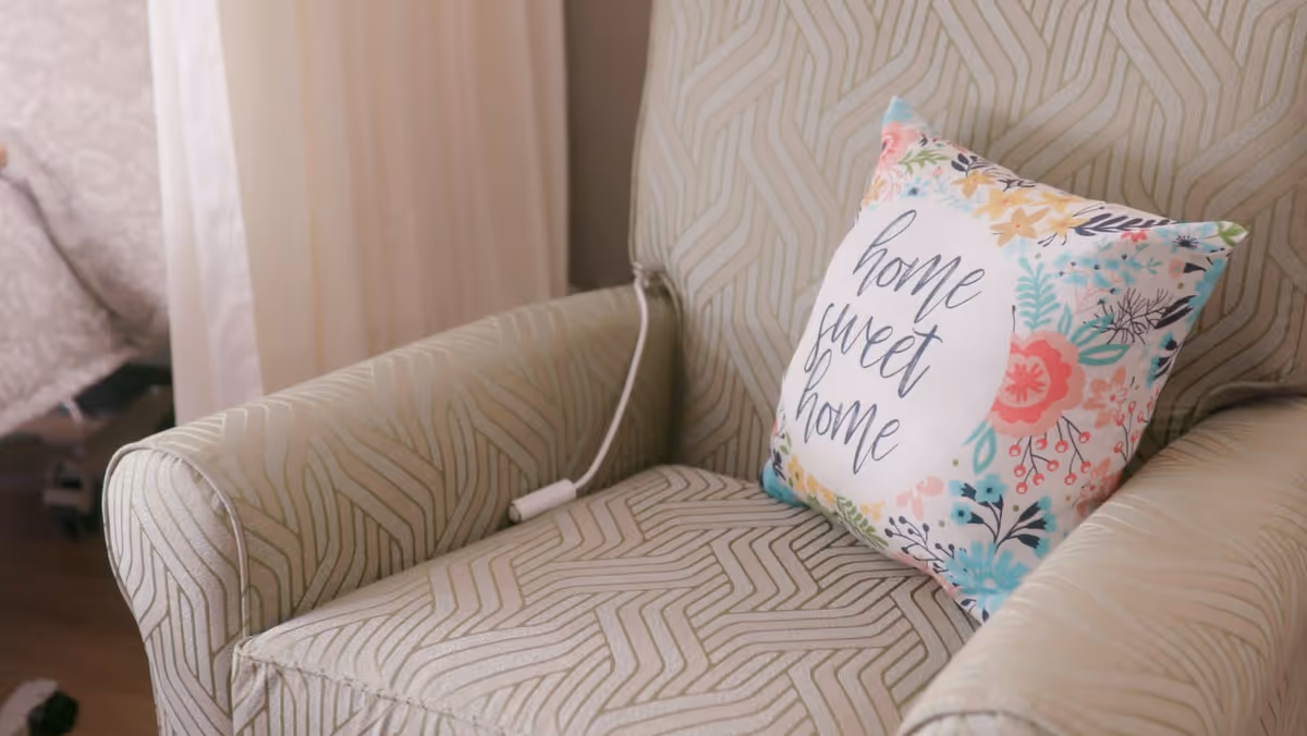 A close-up view of a patterned armchair with a decorative pillow that has floral designs and the words 'home sweet home' written on it. Part of a bed with a patterned bedspread is visible in the background.