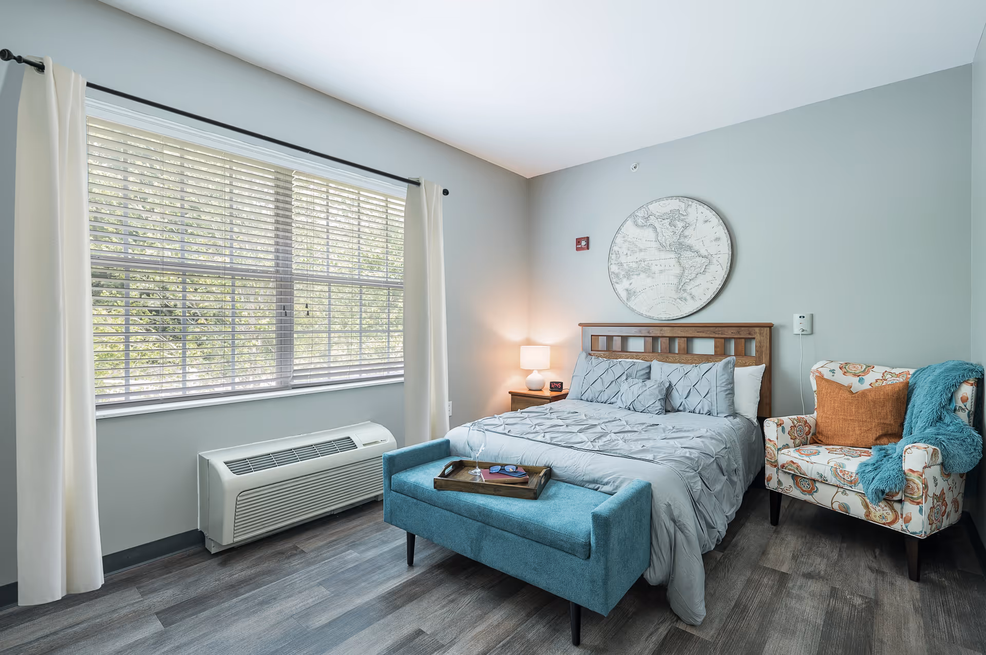 A bright bedroom with a large window covered by white curtains and blinds. The room features a wooden bed with gray bedding and pillows, a small wooden nightstand with a lamp and clock, a blue upholstered bench at the foot of the bed with a tray on top, and a floral-patterned armchair with an orange pillow and a teal throw blanket. The walls are painted light gray, and there is a round map artwork hanging above the bed.