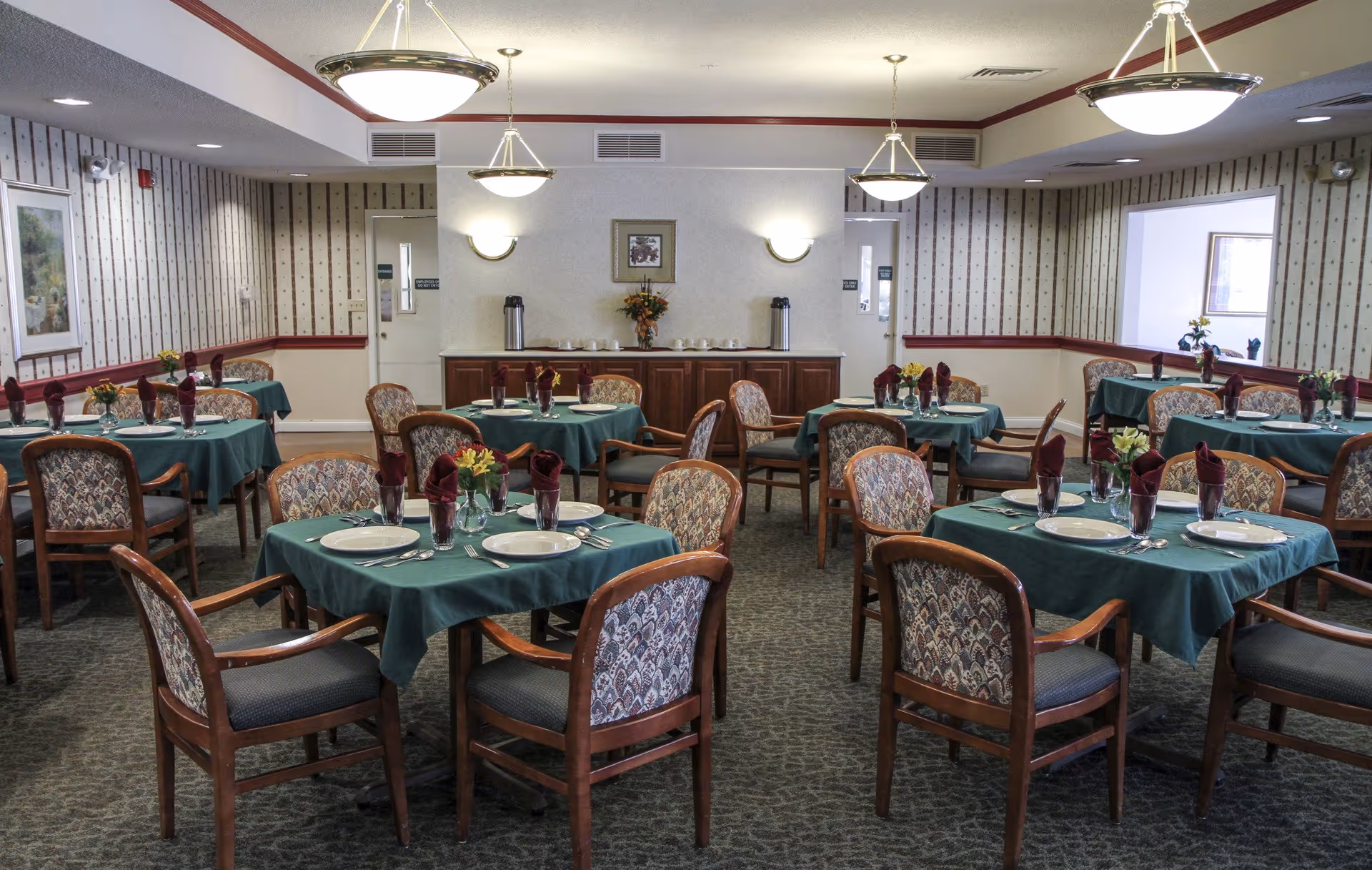 Spacious dining room with multiple round tables covered in green tablecloths, place settings and folded napkins, surrounded by wooden chairs.