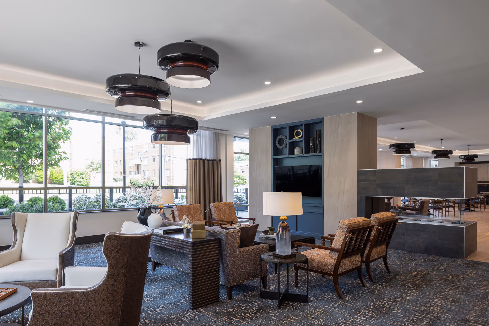 A modern and spacious living room area with large windows letting in natural light. The room features comfortable armchairs and sofas arranged around a central coffee table. There are three large, circular pendant lights hanging from the ceiling. A built-in shelving unit with decorative items and a TV is visible, along with a contemporary fireplace. The room has a patterned carpet and a side table with a lamp.