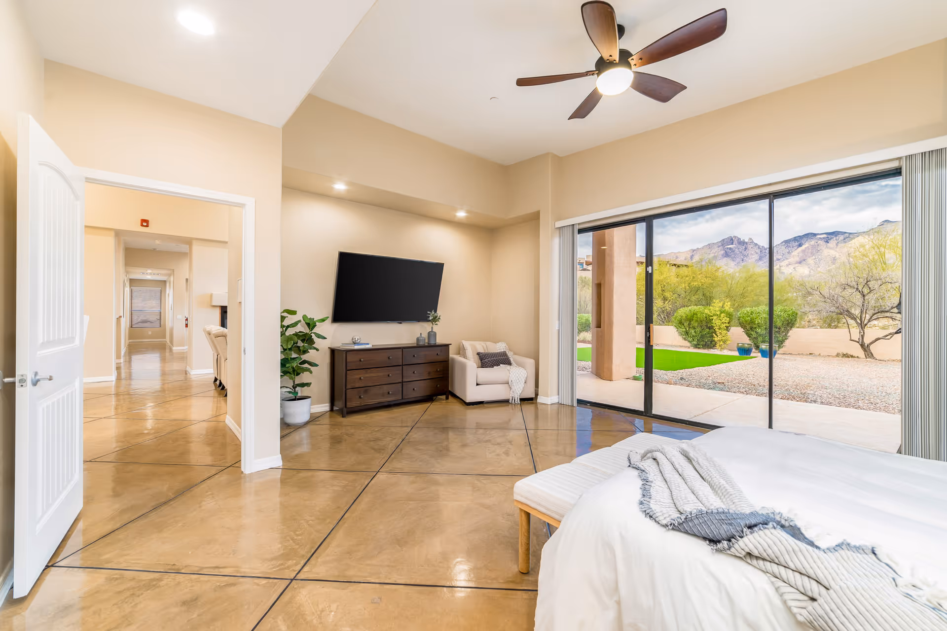 Spacious bedroom with a bed, dresser and wall-mounted TV, seating area, and large sliding glass doors opening to a desert mountain view.