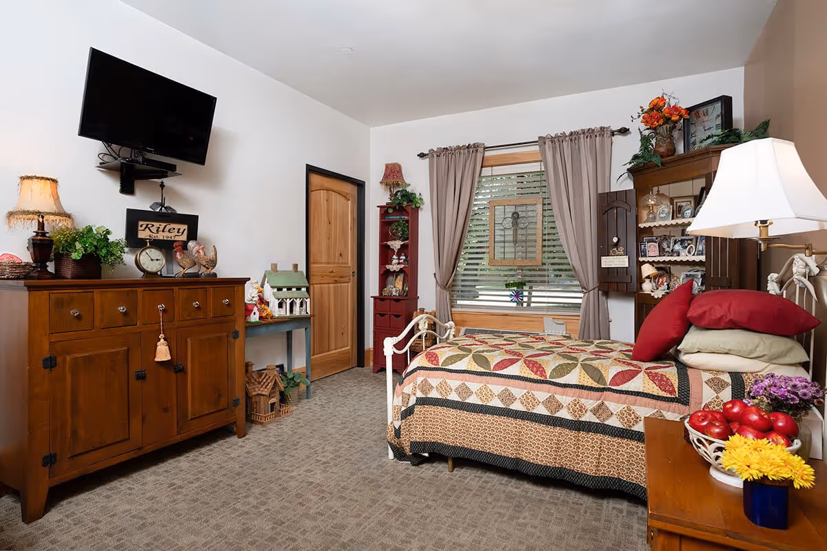 A cozy bedroom in an assisted living facility featuring a bed with a colorful quilt and multiple pillows, a wooden dresser with decorative items including a clock and plants, a wall-mounted TV, a window with beige curtains, and a nightstand with a lamp and flowers.