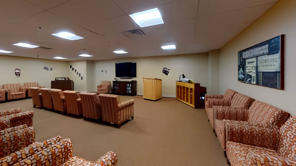 A large room with multiple rows of upholstered chairs and sofas arranged facing a television mounted on the wall. The room has beige walls, a carpeted floor, ceiling lights, a piano, and a podium. A framed artwork is hung on the right wall.