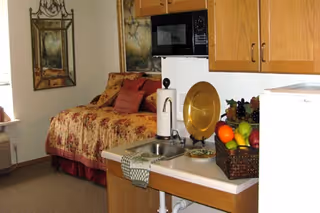 A cozy studio apartment featuring a bed with floral bedding and multiple pillows, a small kitchenette with a sink, microwave, and cabinets, a basket of fresh fruit on the counter, and a framed painting on the wall.