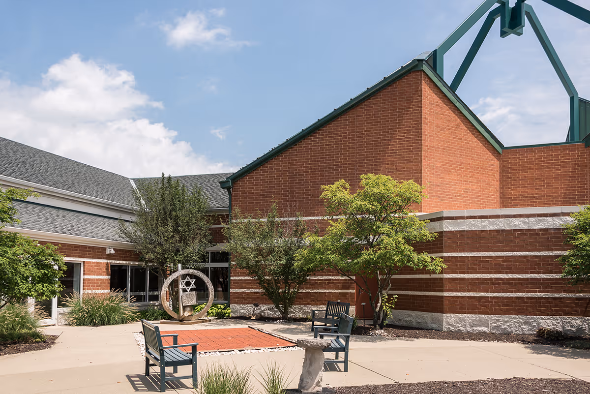 Outdoor courtyard area of a brick building with green-trimmed roof, featuring benches, trees, shrubs, and a circular sculpture with a Star of David symbol under a partly cloudy sky.
