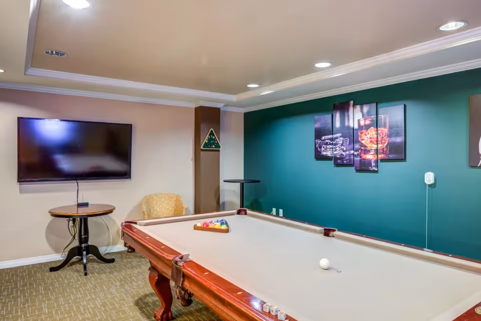 A recreational room with a pool table in the center, a wall-mounted flat screen TV, a small round table, a chair, and a green accent wall with a multi-panel artwork depicting glasses with drinks.