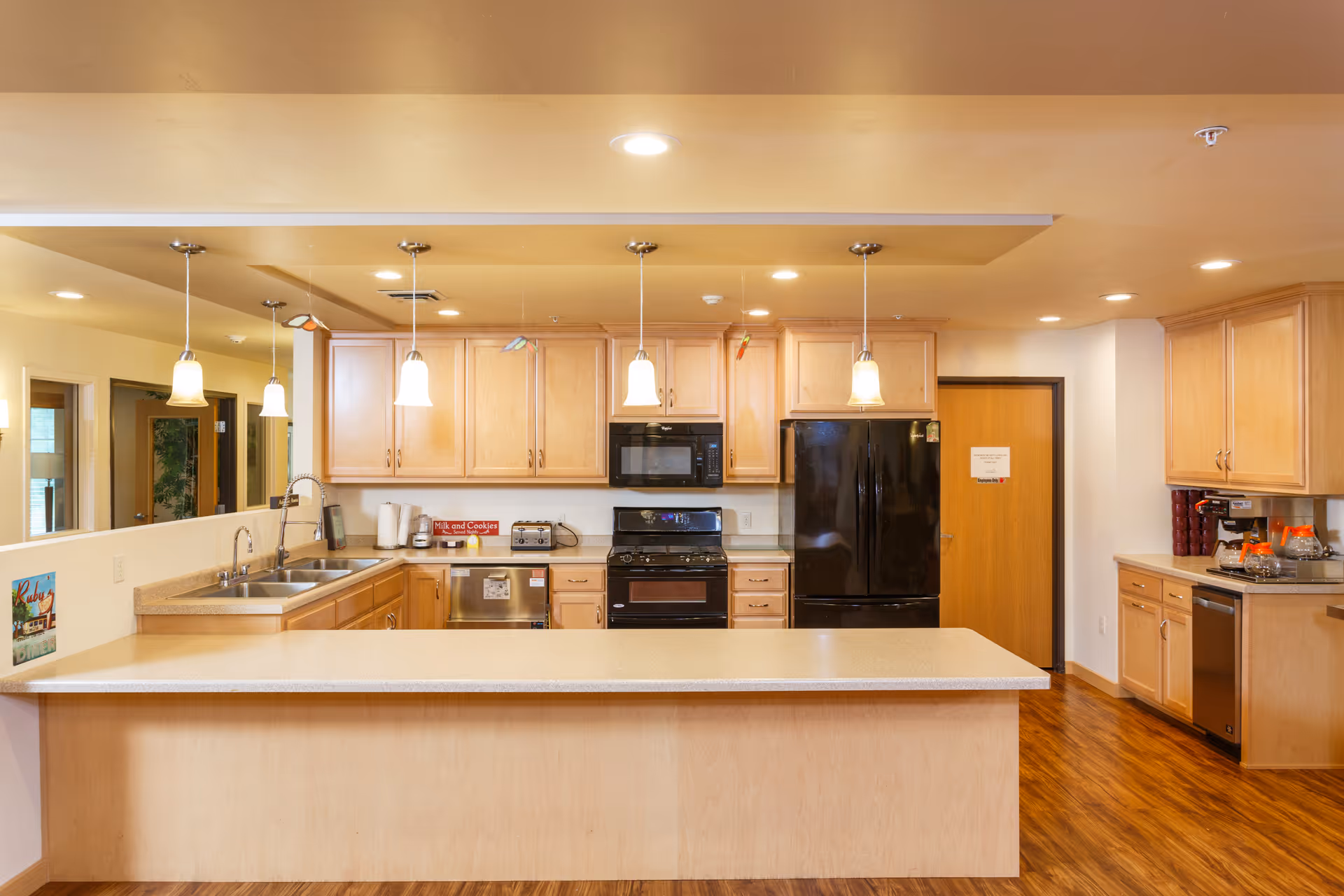 A spacious kitchen area with light wood cabinets, a large island countertop, stainless steel sink, black refrigerator, black microwave, and black oven. The kitchen is well-lit with pendant lights hanging from the ceiling and has a warm wooden floor. There are small decorative items and appliances on the counters.