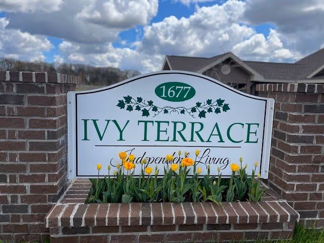 A brick sign with a flower bed in front displaying yellow tulips. The sign reads '1677 IVY TERRACE Independent Living' with decorative ivy leaves above the text. The sky is partly cloudy in the background.
