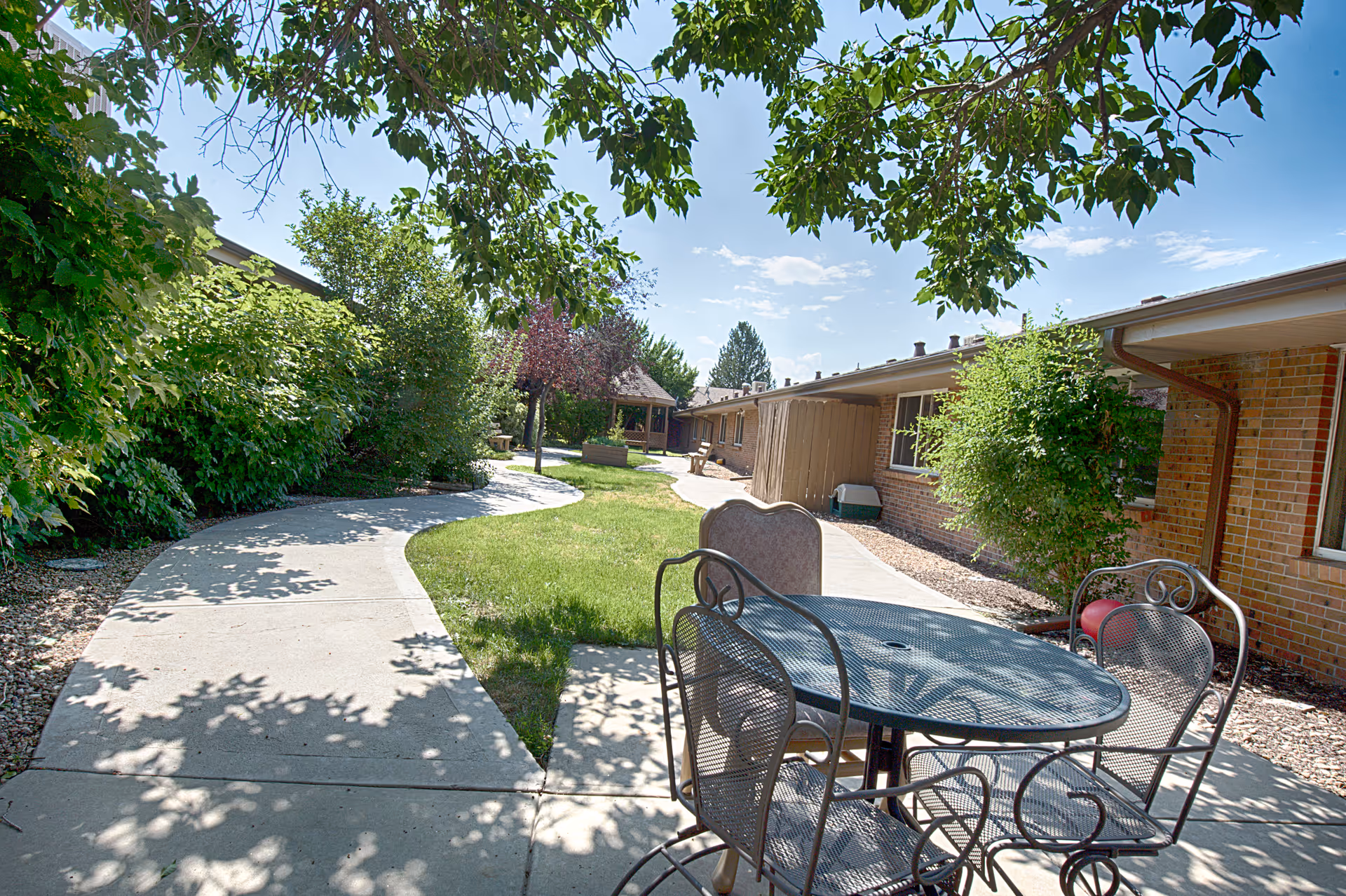 Outdoor patio area at Highland Park Rehabilitation and Care Community with a metal table and chairs on a concrete walkway surrounded by green grass, trees, and bushes under a clear blue sky.