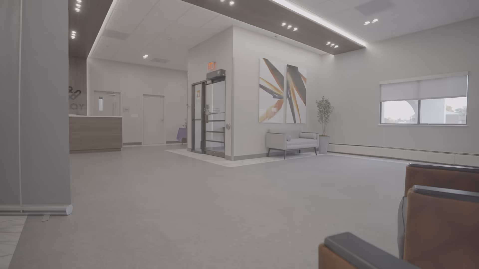 A spacious and modern lobby area with light gray flooring and walls. There is a small gray sofa against the wall with two abstract paintings above it, a potted plant nearby, and a large window with a roller shade. To the left, there is a reception desk and a glass door entrance. The ceiling has recessed lighting and a wood accent panel.
