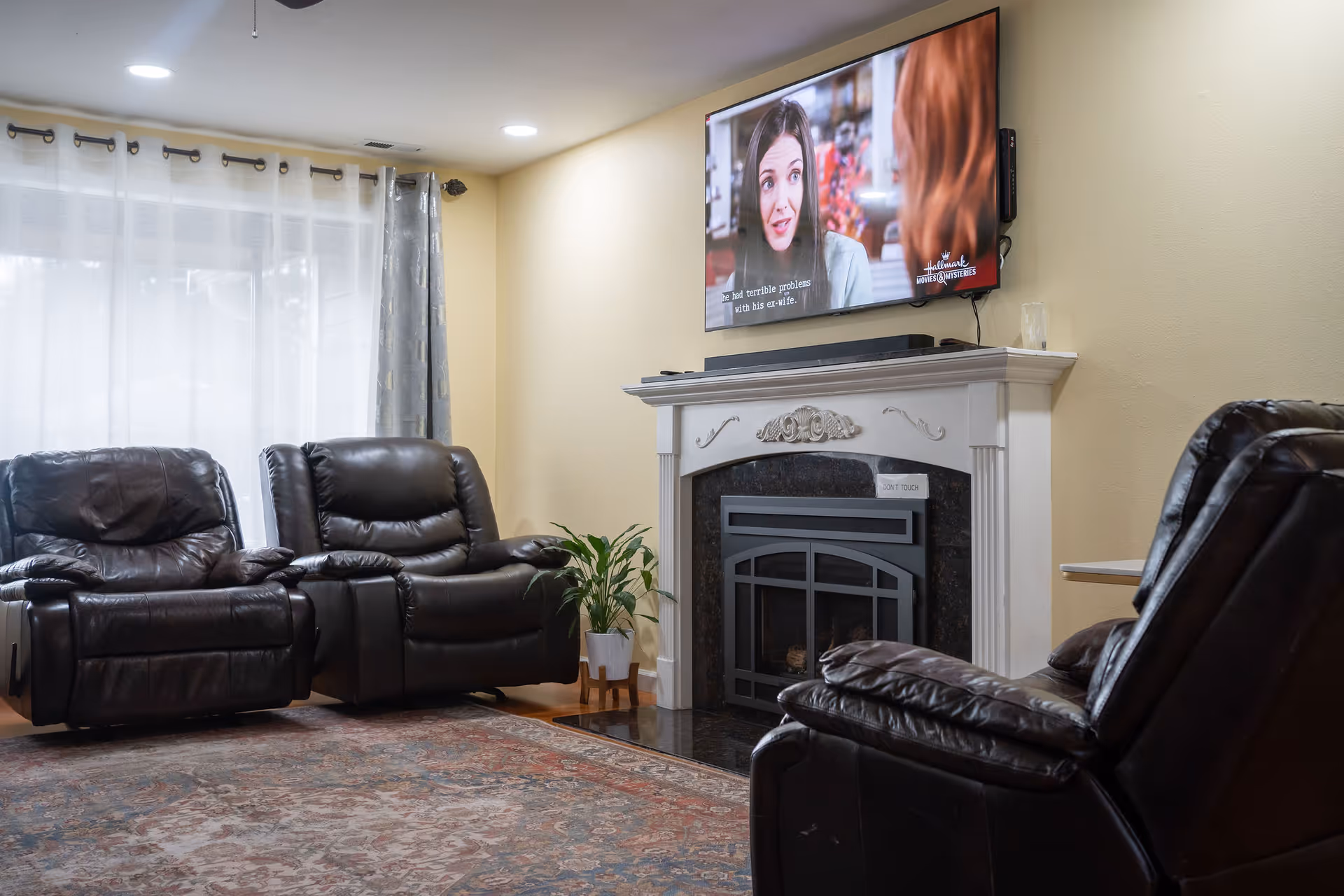 Cozy living room with leather recliners around a decorative fireplace topped by a wall-mounted TV, a patterned rug, and sheer curtains.