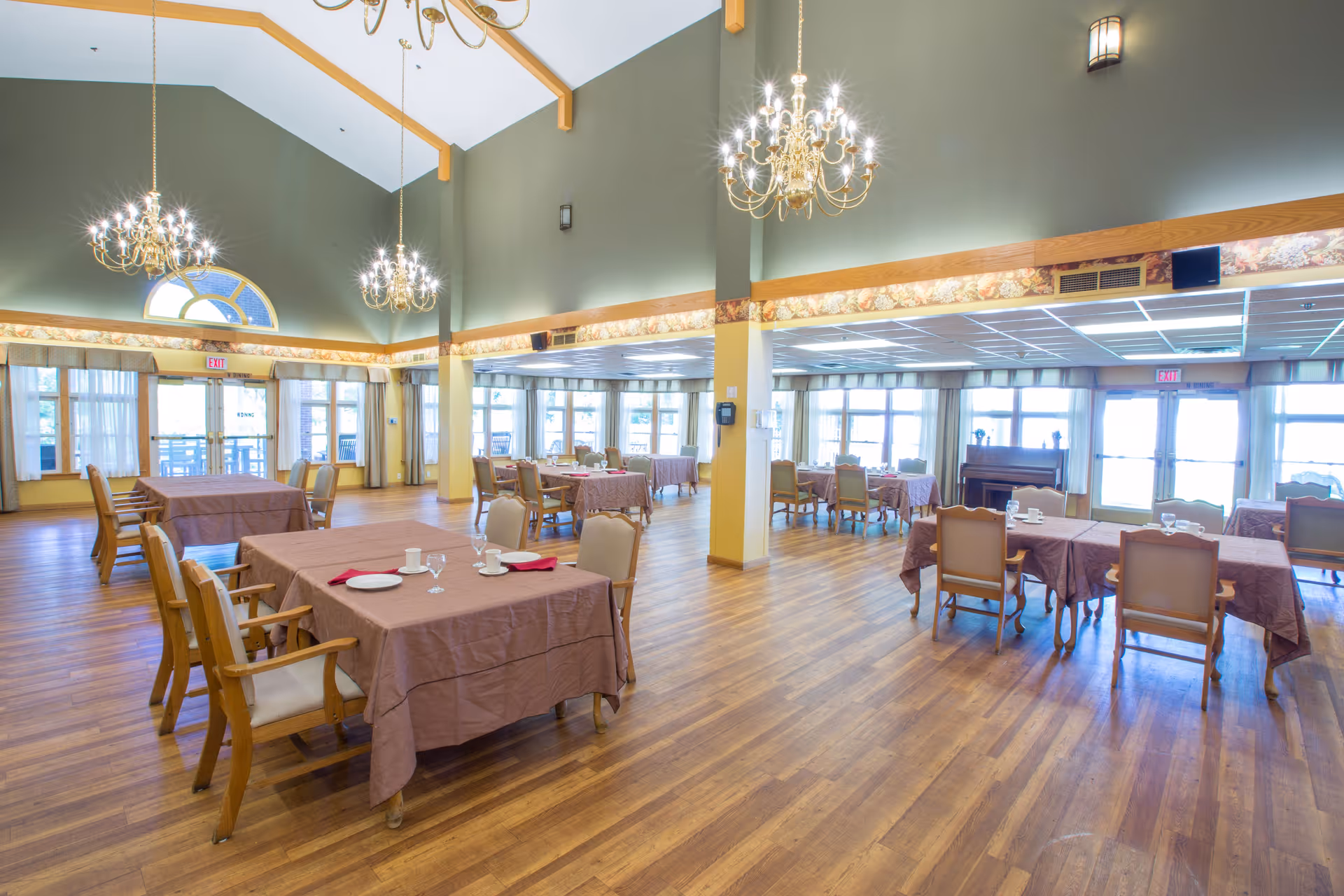 A spacious dining room with wooden floors and multiple tables covered with brown tablecloths. Each table is set with plates, cups, and glasses. The room has high ceilings with chandeliers and large windows letting in natural light. There are chairs arranged around the tables and an exit door at the far end.