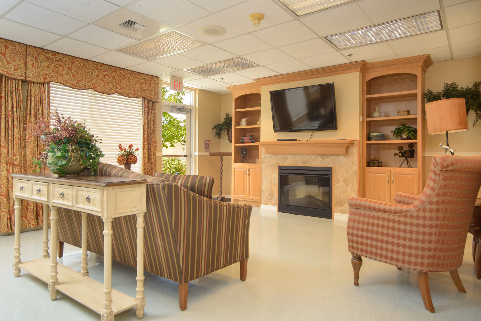 A cozy living room area with a striped sofa, an armchair with a patterned fabric, a wooden console table with a plant on it, a built-in wooden entertainment center with shelves, a flat-screen TV mounted above a fireplace, and large windows with patterned curtains letting in natural light.