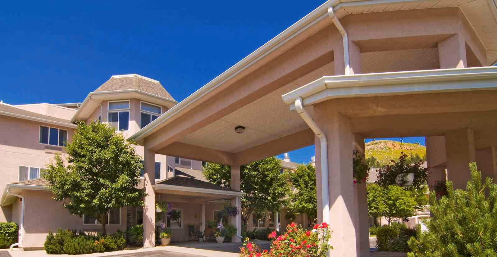Exterior view of Holiday Hills Estates senior living facility showing a covered entrance with columns, surrounding greenery including trees and flowering plants, and a clear blue sky.