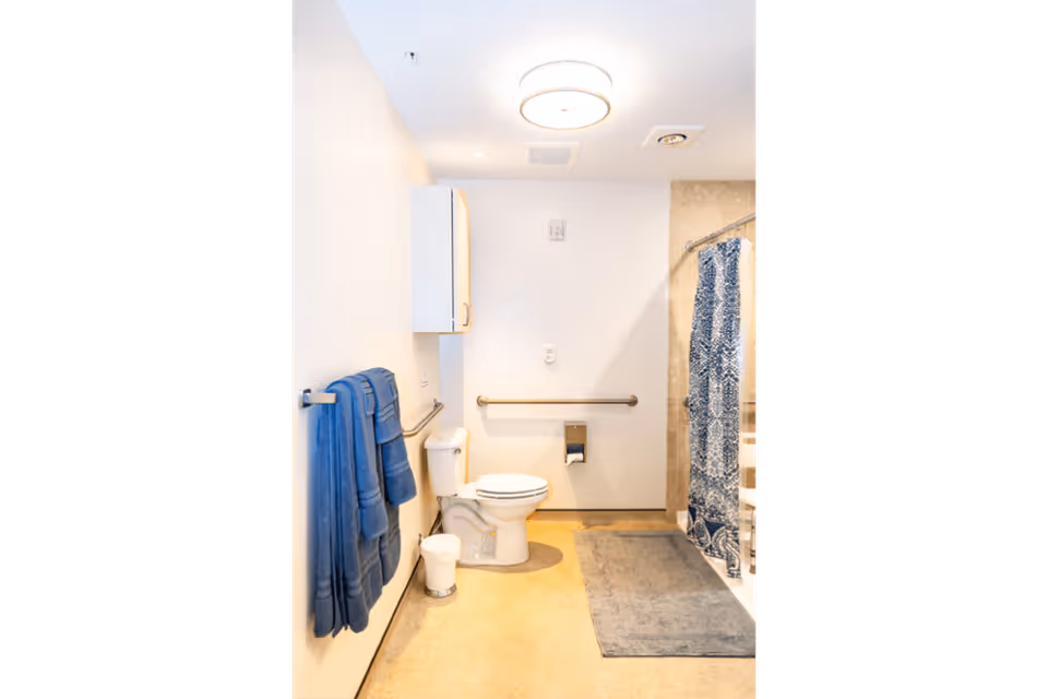 A clean and well-lit bathroom with a toilet, a white cabinet mounted on the wall, blue towels hanging on a towel rack, a gray bath mat on the floor, and a shower area with a blue and white patterned shower curtain.