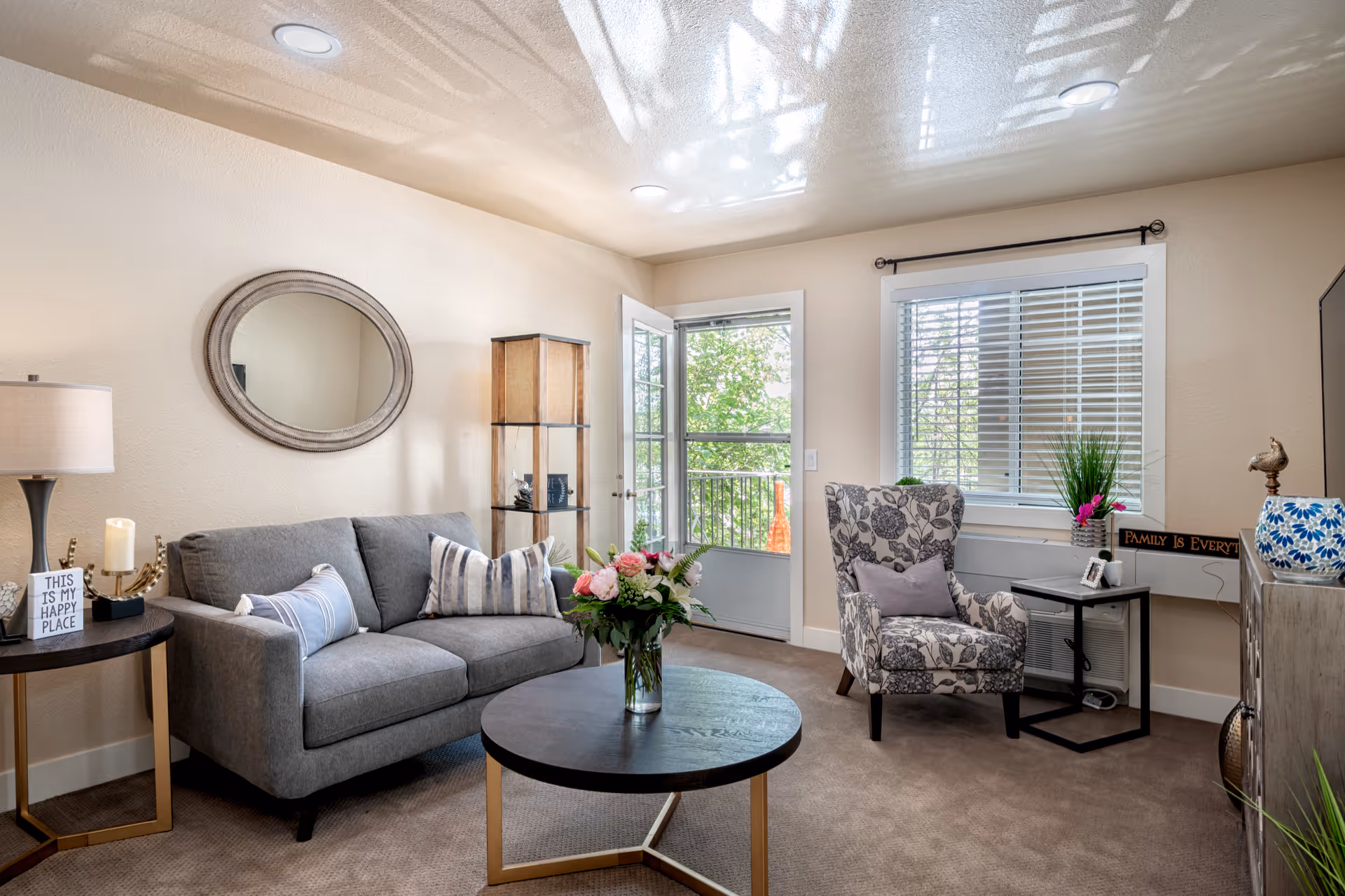A cozy living room with a gray sofa adorned with striped and solid pillows, a round black coffee table with a vase of flowers, a patterned armchair with a lavender pillow, a side table with a lamp and decorative items, a wall mirror, and a glass door leading to a balcony with greenery outside.