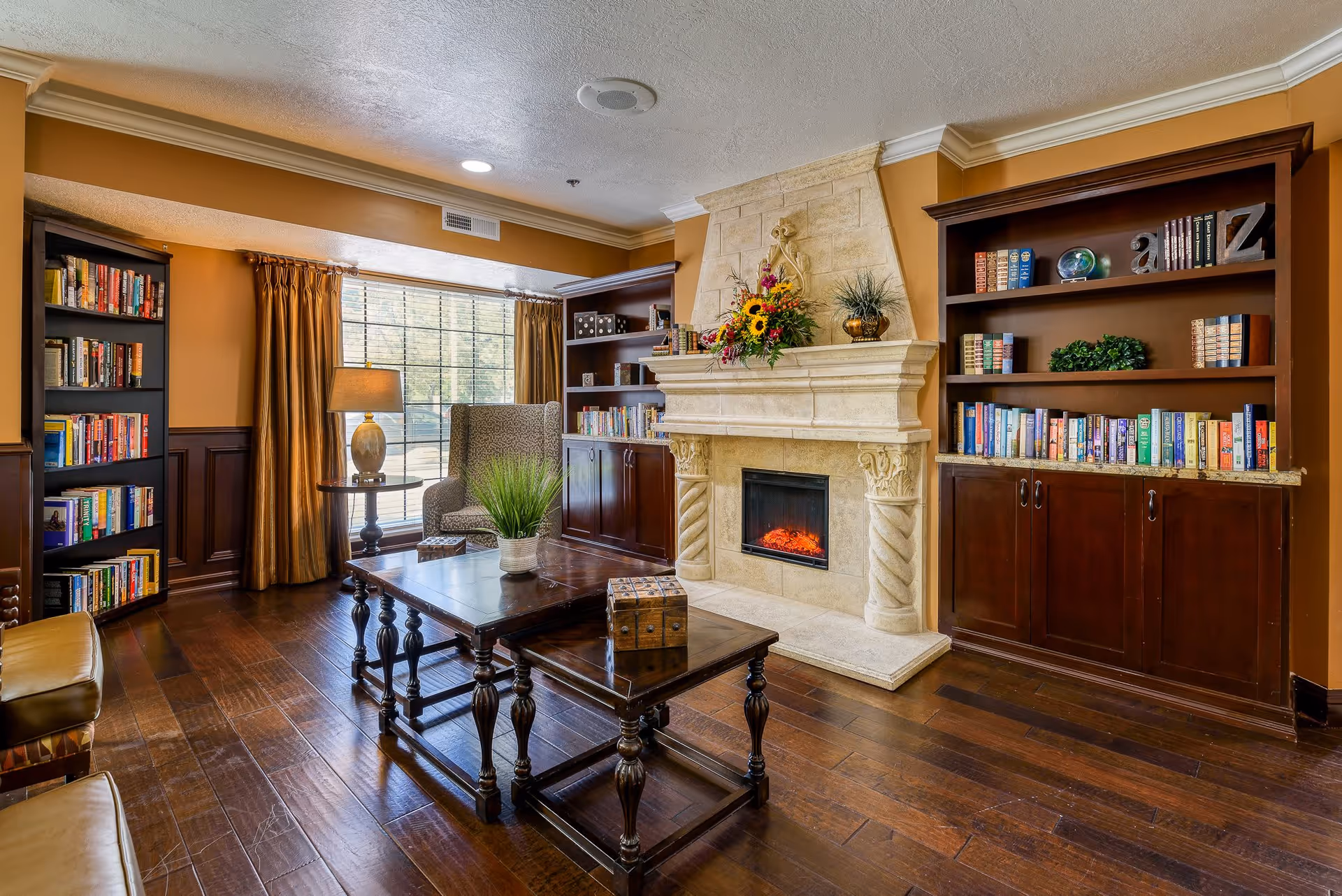 Cozy living room with a stone fireplace flanked by bookshelves, a wooden coffee table, armchair, and a large window with drapes.