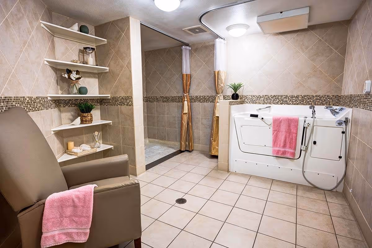 A spacious assisted living bathroom with beige tiled walls and floor. The room features a large walk-in bathtub with a pink towel hanging on its side, a comfortable brown armchair with a pink towel draped over one arm, and built-in white shelves holding decorative items and toiletries. There is also a walk-in shower area with a curtain for privacy.