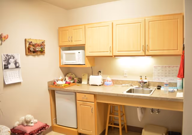 Small kitchenette area with light wood cabinets, a countertop with a sink, a microwave, a toaster, and various kitchen items. There is a small refrigerator under the counter, a wooden stool, and a calendar on the wall. A small white stuffed animal is sitting on a pink cushion on the floor.