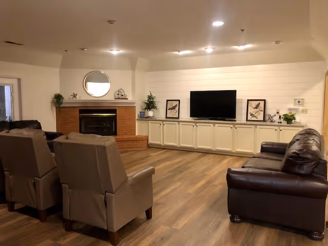 Spacious communal living room with a fireplace, a wall-mounted TV above built-in cabinets, leather sofa and upholstered chairs on wood-look flooring.
