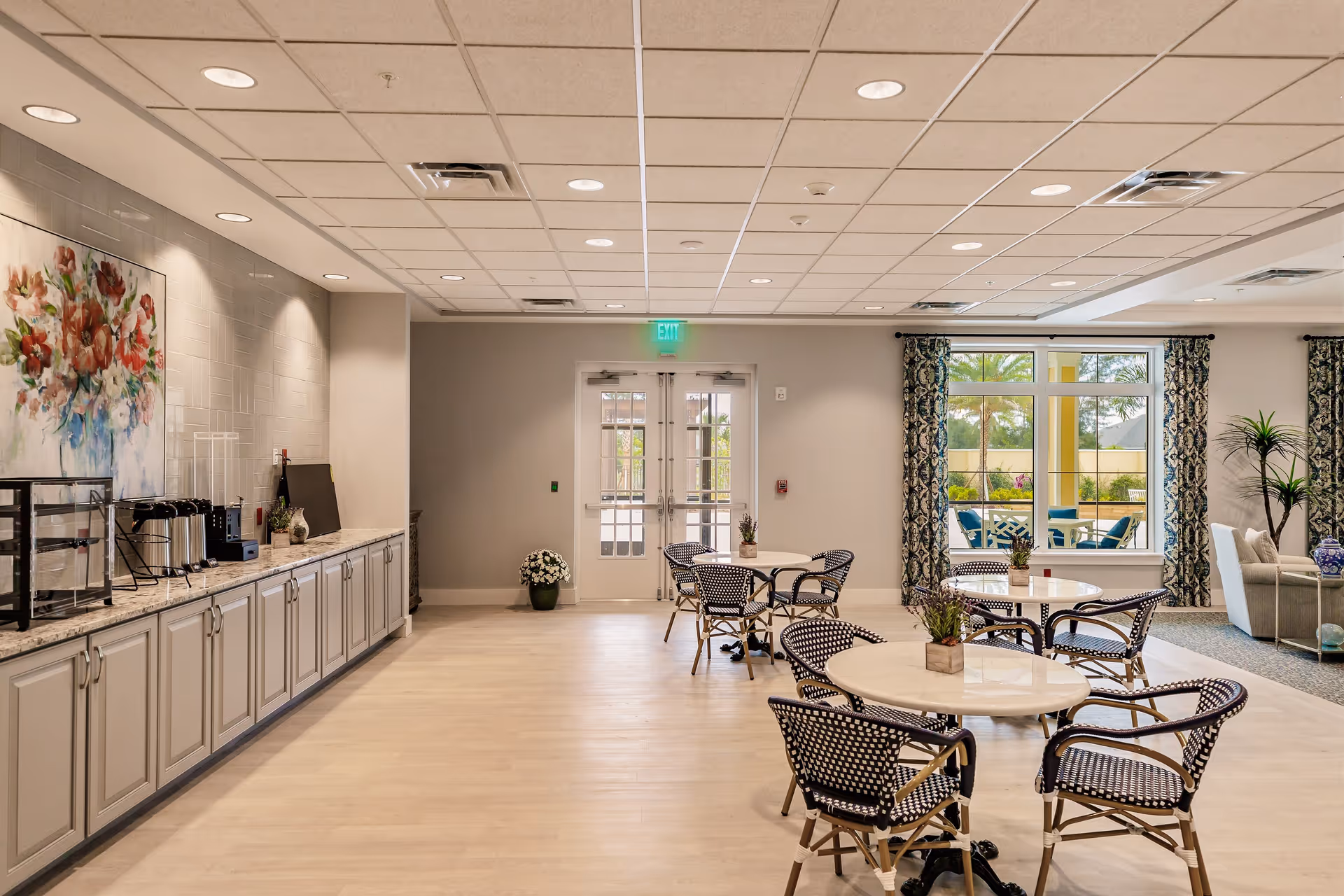 A bright and spacious common area in a senior living facility featuring several round tables with black and white woven chairs. On the left side, there is a long countertop with cabinets underneath, coffee machines, and a floral painting on the wall. Large windows with patterned curtains allow natural light to fill the room, and a glass door leads outside. The room has a light wood floor and a drop ceiling with recessed lighting.
