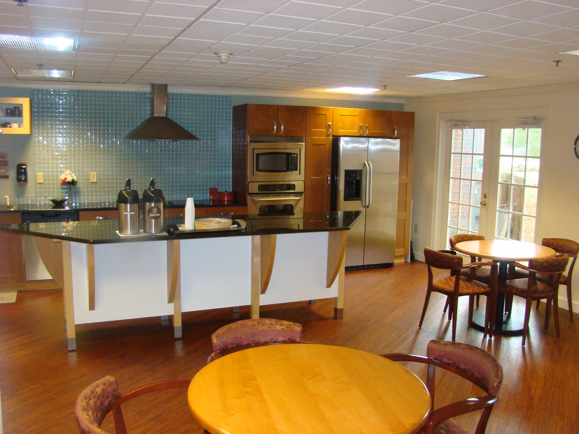 A modern kitchen and dining area with wooden floors, a kitchen island with a black countertop, stainless steel appliances including a refrigerator, oven, and microwave, and round wooden tables with upholstered chairs. There are glass doors leading outside in the background.
