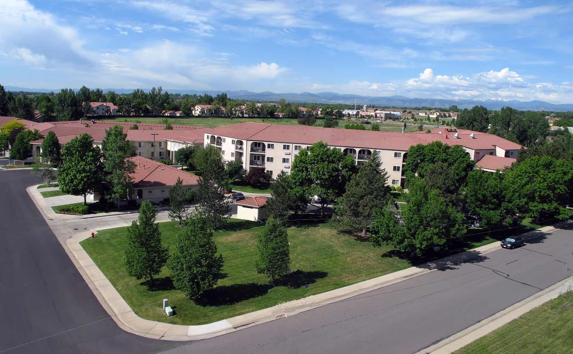 Aerial view of San Marino Retirement Community showing a large multi-story building with a red roof surrounded by green trees and lawns. The building is located at the corner of two paved roads, with mountains visible in the distance under a partly cloudy sky.