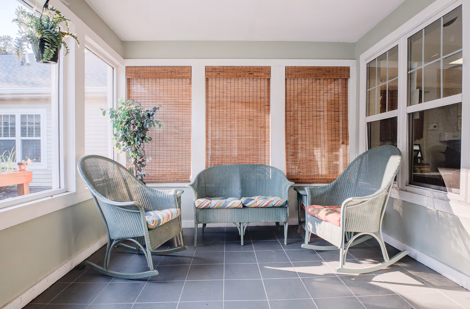 A bright sunroom with three wicker rocking chairs and a wicker loveseat, all with colorful striped cushions. The room has large windows with bamboo blinds and a potted plant in the corner. The floor is tiled in gray.