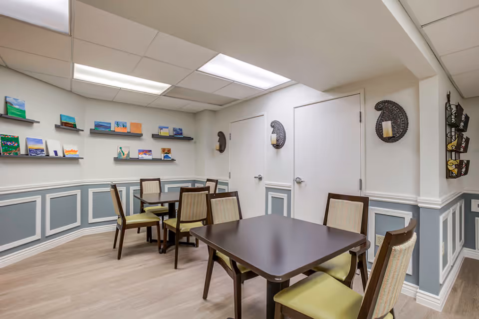 A small room with two dark wood tables and six chairs with light green cushions. The walls are painted white with blue-gray wainscoting and decorated with small paintings on shelves and two decorative wall sconces. The floor is light wood, and the ceiling has fluorescent lighting panels.