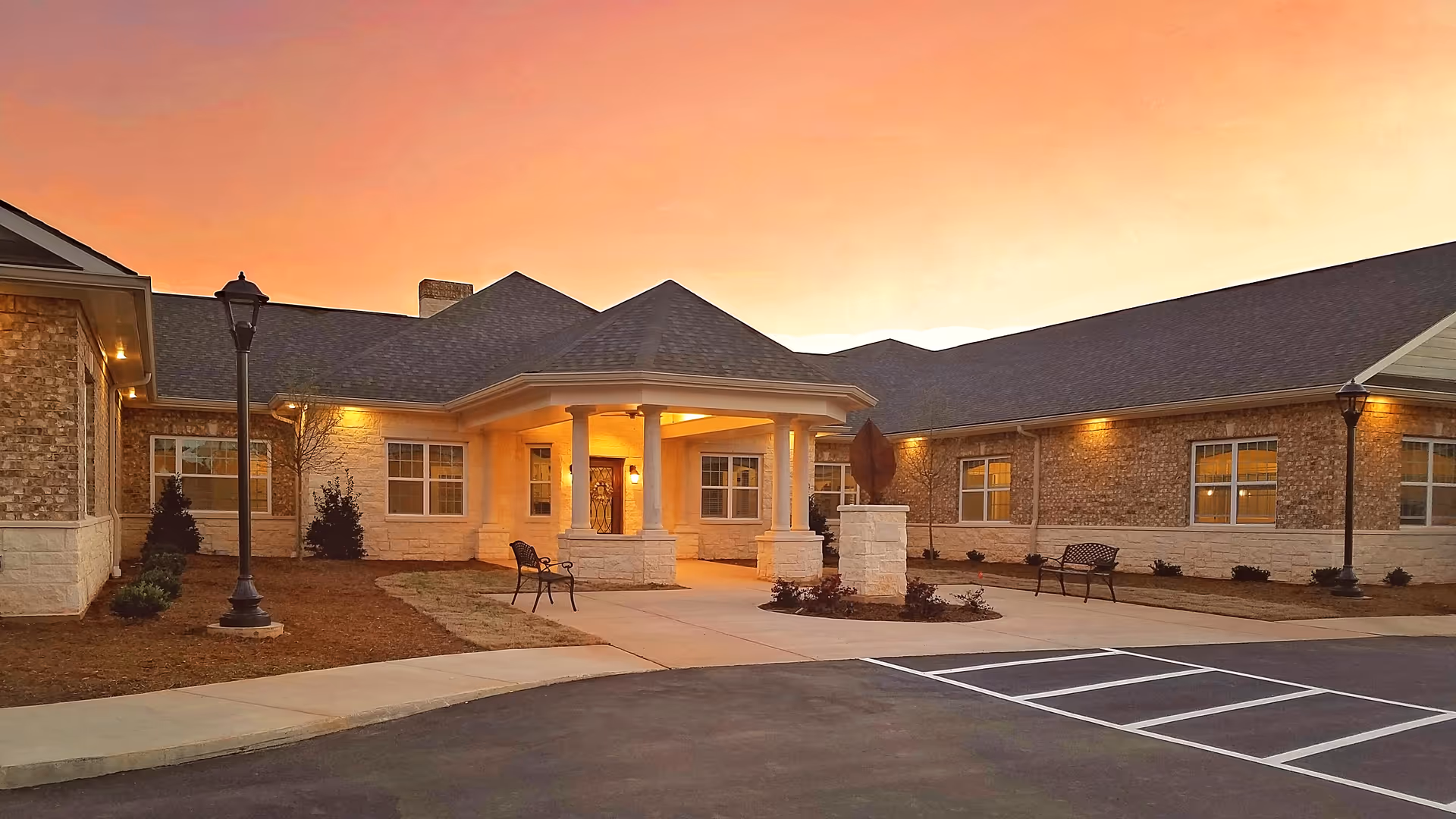 Exterior view of Lavender Hills Fort Mill facility at sunset, showing a single-story building with stone and brick facade, a covered entrance with columns, benches, lamp posts, and a parking area in front.