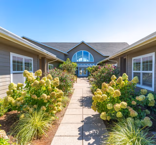 A paved walkway lined with flowering bushes leading to the front entrance of a single-story building with a large arched window.
