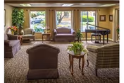 A cozy living room area with multiple upholstered chairs and sofas arranged around small wooden tables. Large windows let in natural light and offer a view of greenery outside. A grand piano is positioned near the windows, and the room is decorated with plants and framed pictures on the walls.