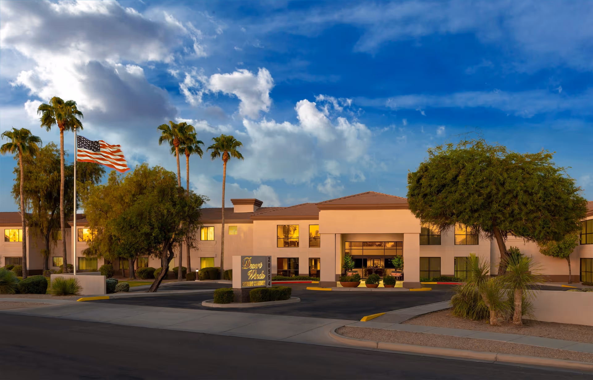 Exterior view of Desert Winds Assisted Living facility during sunset with palm trees, an American flag on a flagpole, and a clear sky with scattered clouds. The building has a beige facade with large windows and a covered entrance.