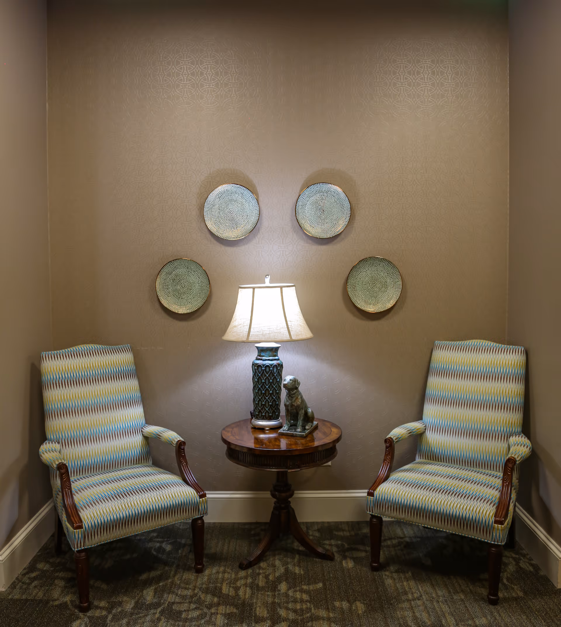 Two upholstered armchairs flank a small round wooden table topped with a lamp and statuette beneath decorative wall plates in a cozy seating nook.