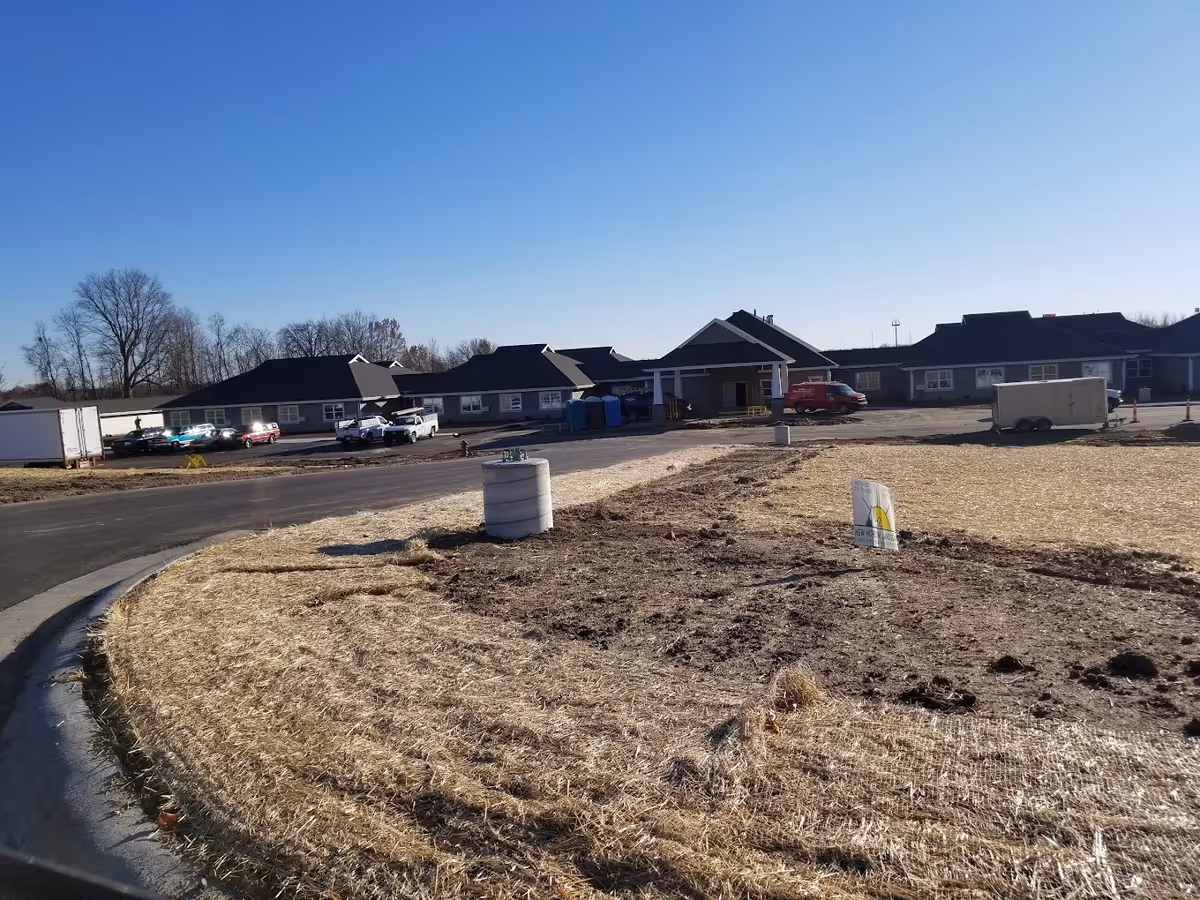 Front exterior of a senior living facility under construction with parked vehicles, trailers, and straw-covered ground under a clear blue sky.
