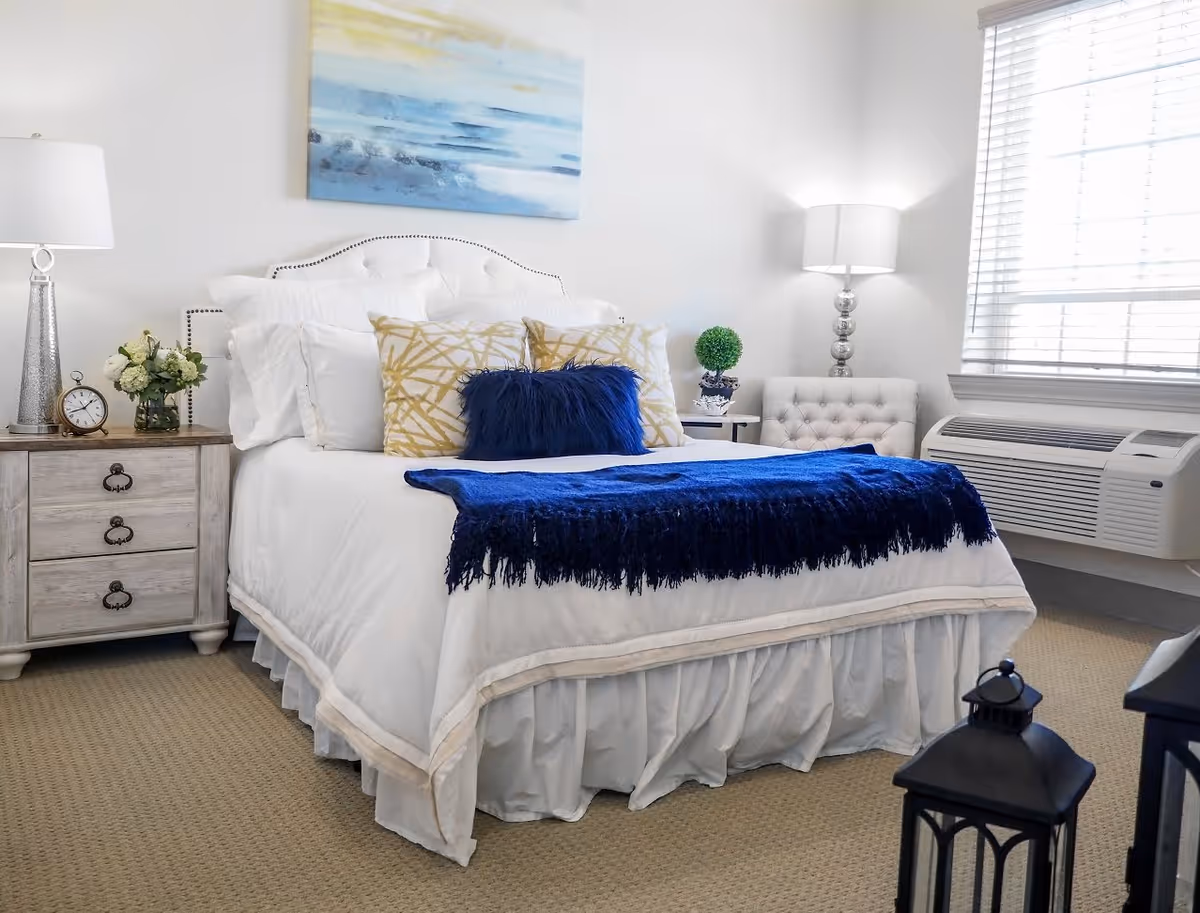 A neatly made bed with white bedding, decorative pillows in white, gold, and blue, and a blue throw blanket. The room features a nightstand with a lamp, clock, and flowers, a tufted chair, a small side table with a plant, a window with blinds, and a wall-mounted air conditioning unit. A painting with blue and yellow tones hangs above the bed.