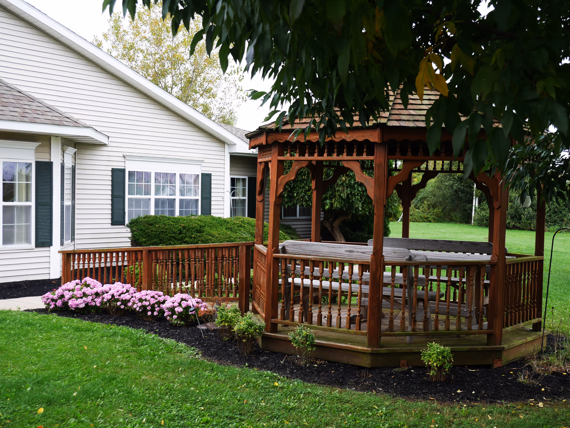 A wooden gazebo with benches inside situated on a grassy lawn next to a white building with green shutters. There are pink flowers and bushes planted along the edge of the building and gazebo, with trees and greenery in the background.