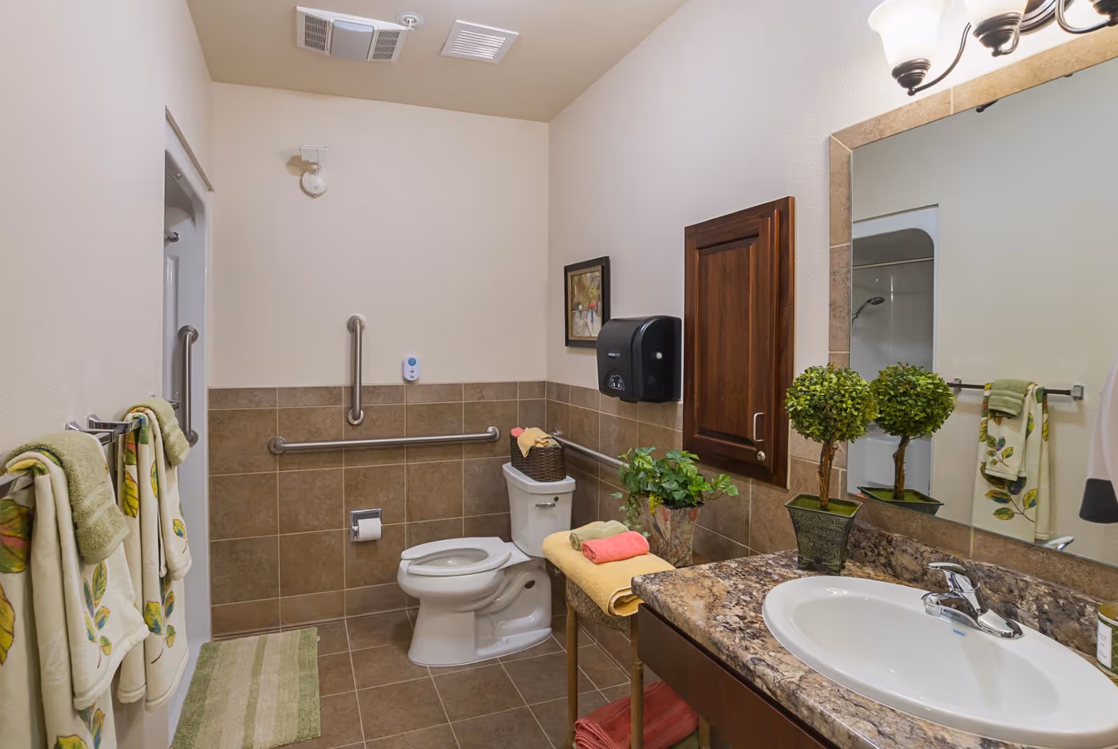 A clean and well-lit bathroom with beige tiled walls and floor. The bathroom features a white toilet with grab bars on the walls for accessibility, a granite countertop with a white sink, a large mirror, and a wooden cabinet mounted on the wall. There are decorative plants on the countertop and a small table holding folded towels. Towels with leaf patterns hang on a rack next to the shower area.